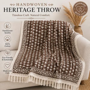 May include: A handwoven throw blanket featuring a dark brown base with a repeating white floral pattern and a decorative border. The throw has cream-coloured fringe along the edges and includes text reading Handwoven Heritage Throw.