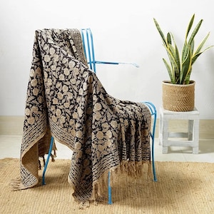 May include: A black and tan floral patterned throw blanket with fringe is draped over a light blue metal chair. The blanket has a repeating floral design and a decorative border. A potted plant sits on a small white table.