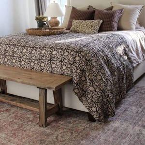 Könnte beinhalten: Ein weißes Bett mit einer schwarz-weißen gemusterten Bettdecke und einer Holzbank am Fußende des Bettes. Das Bett hat ein weißes Kopfteil und einen braunen Teppich auf dem Boden.