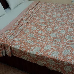 Puede incluir: Una colcha de color coral con un estampado floral en blanco y gris cubre una cama. La colcha está drapeada sobre la cama, mostrando el intrincado diseño. La cama tiene almohadas blancas y un borde rosa claro.