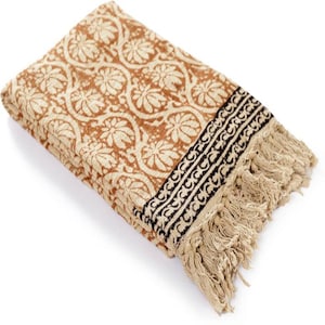 May include: Folded beige and brown patterned textile with a floral design. The textile has a black border with a repeating pattern and fringed edges. The textile is folded and appears to be made of a soft material.