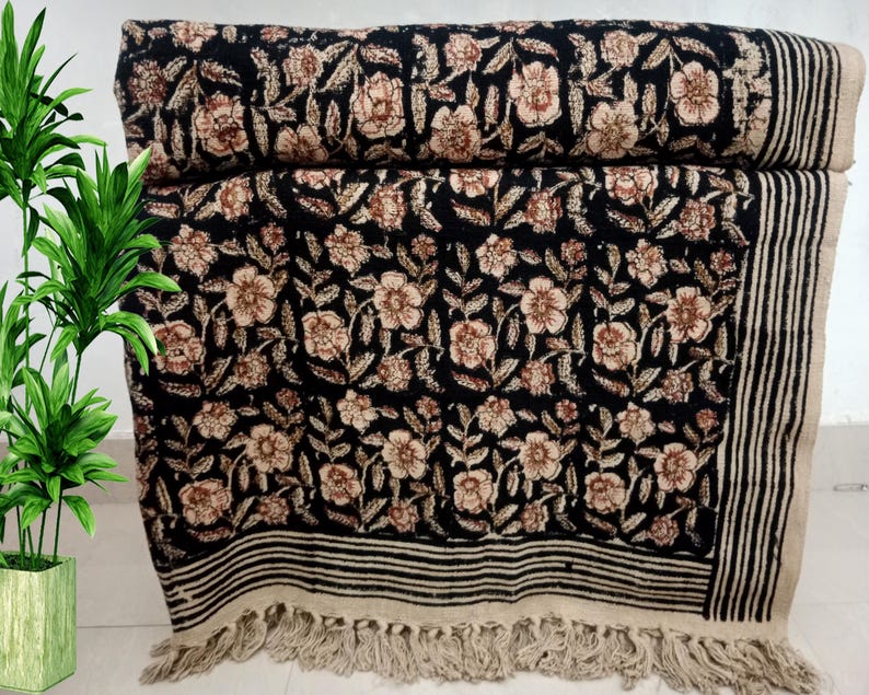 May include: A black floral patterned textile, possibly a throw blanket or rug, with a repeating design of beige flowers and leaves. The edges feature black and white stripes and a fringe. The textile is folded and stacked.