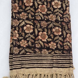 May include: Folded dark brown floral blanket with a beige fringe. The blanket features a repeating pattern of flowers and leaves in beige and red. The bottom edge has horizontal black stripes and a beige fringe.