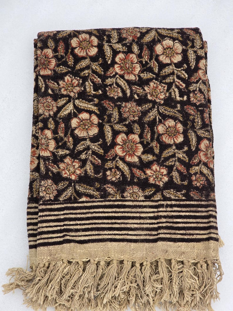 May include: Folded dark brown floral blanket with a beige fringe. The blanket features a repeating pattern of flowers and leaves in beige and red. The bottom edge has horizontal black stripes and a beige fringe.