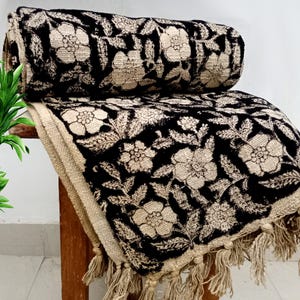 Könnte beinhalten: Eine schwarz-beige Decke mit floralem Muster und Fransen. Die Decke ist über einer Holzoberfläche drapiert, wobei ein aufgerollter Teil sichtbar ist. Das florale Design zeigt große Blumen und Blätter.