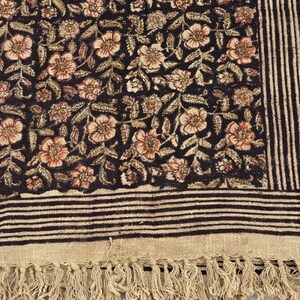 May include: A dark textile with a floral pattern in shades of brown, beige, and red. The fabric features a striped border and a fringed edge. The floral design is densely packed, creating a rich, textured appearance.