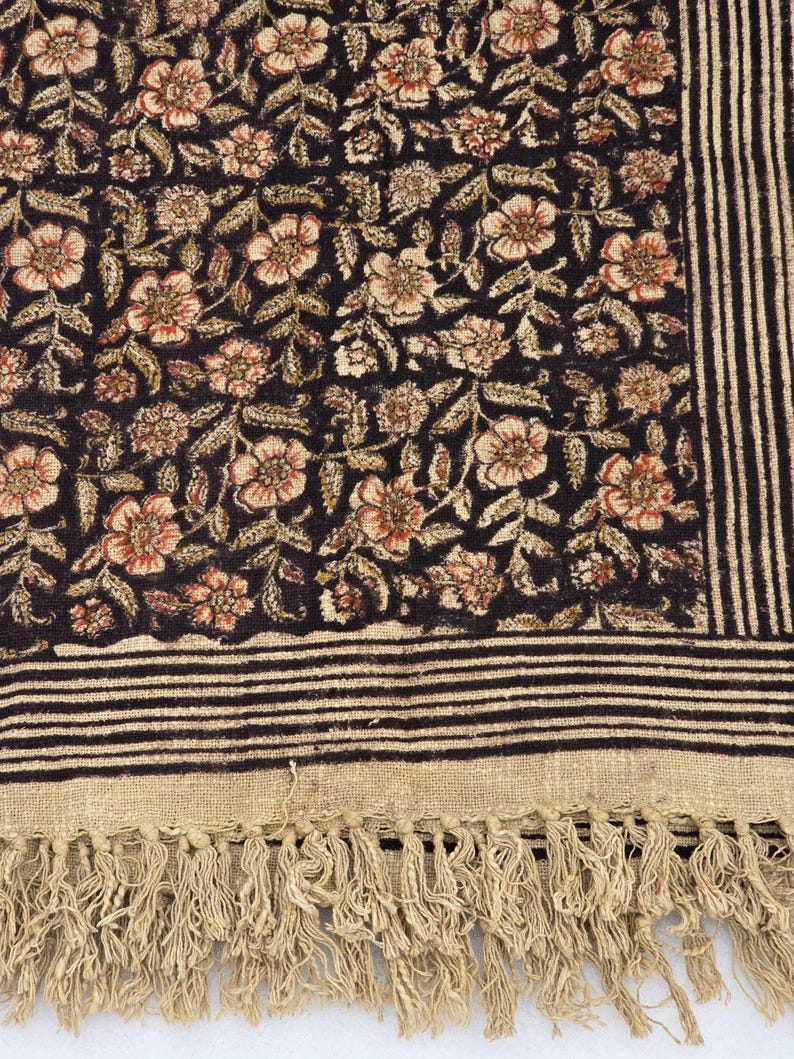 May include: A dark textile with a floral pattern in shades of brown, beige, and red. The fabric features a striped border and a fringed edge. The floral design is densely packed, creating a rich, textured appearance.