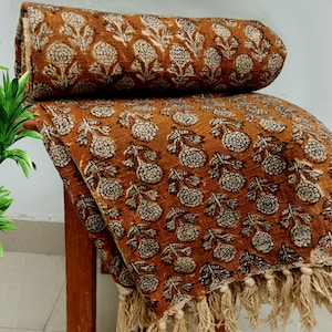 Puede incluir: Una manta de tiro de color marrón y blanco con estampado floral y borde de flecos. La manta está doblada y drapeada sobre una silla de madera.