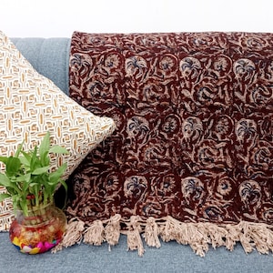 Puede incluir: Una manta decorativa burdeos con estampado floral y flecos color crema. También se ven una almohada estampada y un pequeño jarrón de cristal con plantas. La manta mide unos 150 cm de largo.