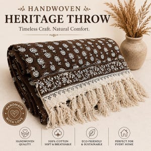 Pode incluir: Uma manta castanha escura dobrada com um padrão floral branco, uma borda decorativa com motivo de videira e borlas de franjas cor de creme. O texto HANDWOVEN HERITAGE THROW Timeless Craft. Natural Comfort. é visível acima da manta.