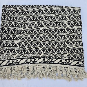 May include: A rectangular textile featuring a repeating black fan-shaped pattern on an off-white background. The bottom edge displays a horizontal border with a black floral motif, finished with a row of off-white knotted fringe tassels.