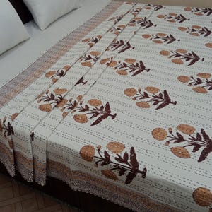 May include: White bedspread with a repeating pattern of brown floral motifs and dark brown leaves. The bedspread has a striped design and a brown border. The bedding is on a bed with white pillows.