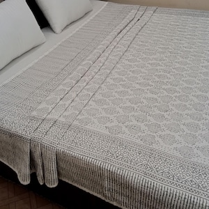 May include: A white and grey patterned bedspread with two white pillows. The bedspread features a repeating floral design and a striped border. The bedding is neatly arranged on a bed.