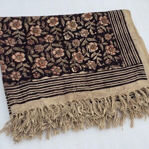 May include: A rectangular textile with a floral pattern in shades of brown and beige on a black background. The border features horizontal stripes and fringe. The textile is folded.