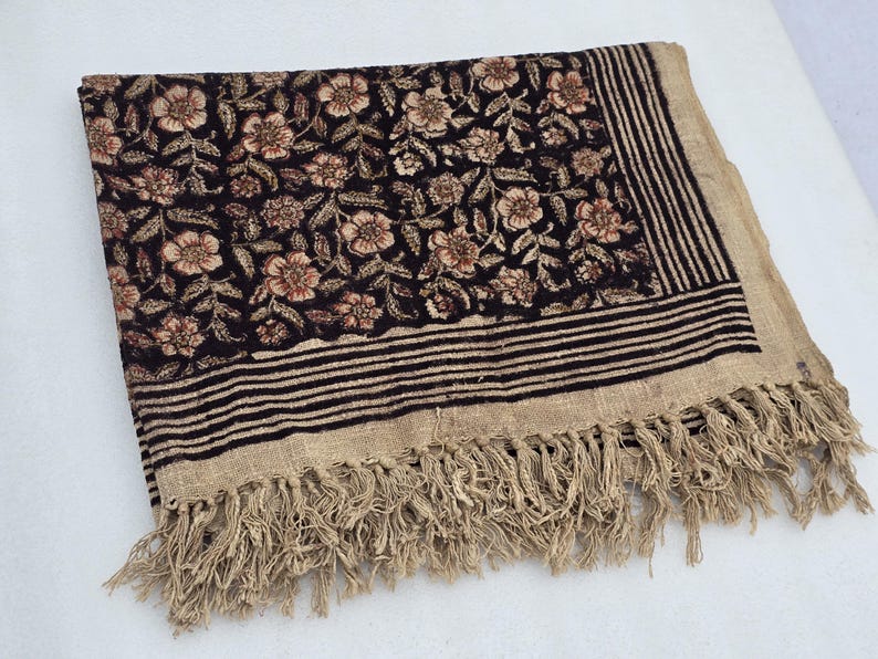 May include: A rectangular textile with a floral pattern in shades of brown and beige on a black background. The border features horizontal stripes and fringe. The textile is folded.