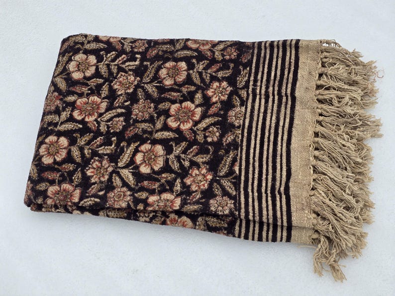 May include: A folded textile with a floral pattern in shades of brown and beige on a black background. The edge features vertical stripes and a fringe in a neutral colour. The textile appears to be a blanket or throw.