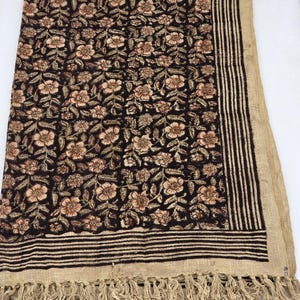 May include: A dark brown floral patterned textile with a beige fringe. The textile features a repeating pattern of flowers in shades of beige and brown. The border has vertical stripes and a fringe.