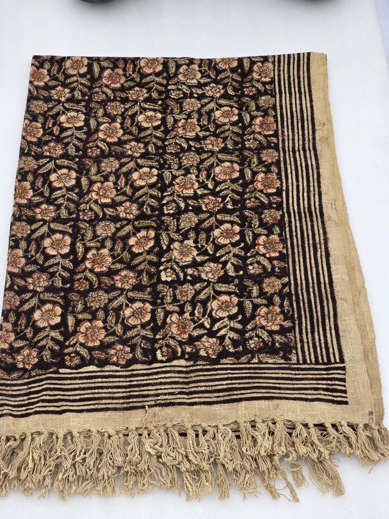 May include: A dark brown floral patterned textile with a beige fringe. The textile features a repeating pattern of flowers in shades of beige and brown. The border has vertical stripes and a fringe.