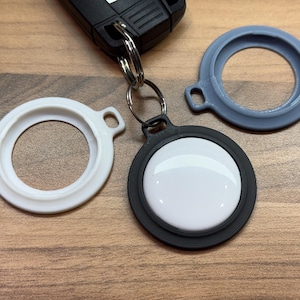 Op de afbeelding: Een verzameling sleutelhangers en beschermhoezen voor een elektronisch apparaat. De afbeelding toont een zwart-wit apparaat met een zwarte hoes bevestigd aan een sleutelhanger, samen met twee extra hoezen in wit en grijs. De items liggen op een houten oppervlak.