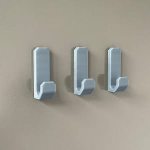 Könnte beinhalten: Drei hellgraue Wandhaken mit rechteckiger Oberseite und gebogener Hakenform. Die Haken sind gleichmäßig angeordnet und an einer neutralfarbenen Wand montiert, was ein schlichtes und funktionales Design bietet.