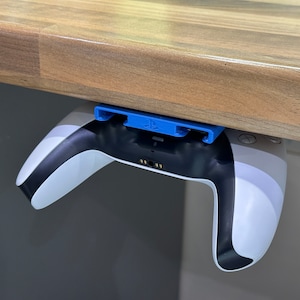 Può includere: Un controller per videogiochi bianco e nero è montato sotto un tavolo di legno con un supporto in plastica blu. Il controller è capovolto con i pulsanti rivolti verso il tavolo.