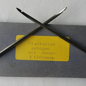 Könnte beinhalten: Zwei silberfarbene, gebogene, hakenförmige Werkzeuge mit einem gelben Etikett, auf dem "Plattahlen gebogen mit Haken 1.00mm" steht.