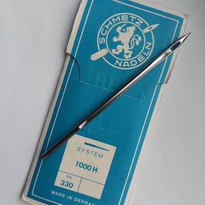 May include: A single sewing needle rests on a blue and white Schmetz needle package. The package features a logo with a lion and the text "SCHMETZ NADELN". The package also includes the text "SYSTEM 1000 H" and "Nm 330".