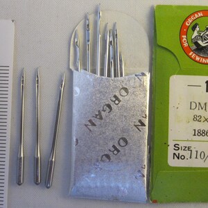 Peut inclure: Un paquet de 10 aiguilles à coudre pour machine à coudre en argent dans un sachet en papier argenté et blanc. Le paquet est étiqueté "ORGAN" et "NY". Les aiguilles sont également présentées individuellement à côté d'une règle. Le paquet est étiqueté "ORGAN NEEDLES FOR SEWING MACHINES" et "SIZE No. 110/18".