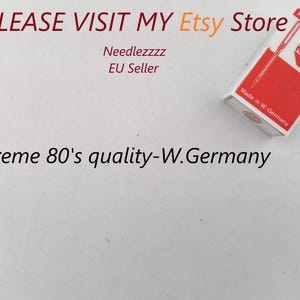 Puede incluir: Una caja roja y blanca con el texto "Rhein Nadel" y "Precision Quality" impreso en ella. La caja contiene una aguja de coser. El texto "Made in W-Germany" está impreso en la parte inferior de la caja. El texto "Supreme 80's quality-W.Germany" está impreso debajo de la caja.