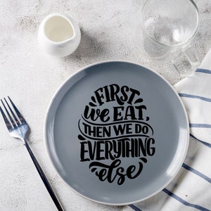 以下が含まれることがあります： 黒い文字で「First we eat, then we do everything else」と書かれた灰色のセラミックプレート。プレートは白い表面に置かれ、隣には白いクリーマーと透明なグラスがある。