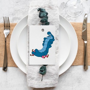 May include: A white place card with a blue caterpillar illustration. The caterpillar is sitting on a mushroom and has a surprised expression. The place card is on a white plate with a gray linen napkin.