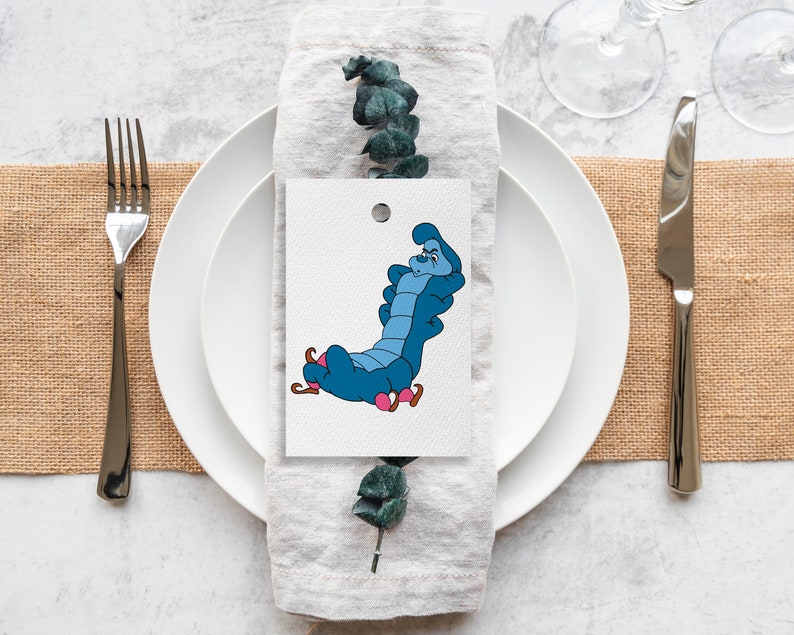 May include: A white place card with a blue caterpillar illustration. The caterpillar is sitting on a mushroom and has a surprised expression. The place card is on a white plate with a gray linen napkin.