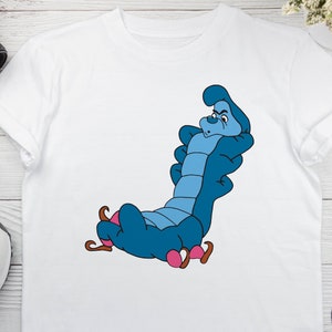 May include: White t-shirt with a blue caterpillar character from Alice in Wonderland. The caterpillar is sitting on a pink mushroom and has a grumpy expression.