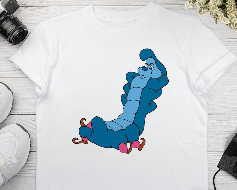 May include: White t-shirt with a blue caterpillar character from Alice in Wonderland. The caterpillar is sitting on a pink mushroom and has a grumpy expression.