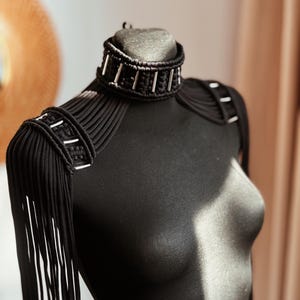 Peut inclure: Un collier ras de cou noir avec des accents argent&eacute;s et des bretelles assorties avec des franges. Le collier et les bretelles sont en cordon tiss&eacute;.