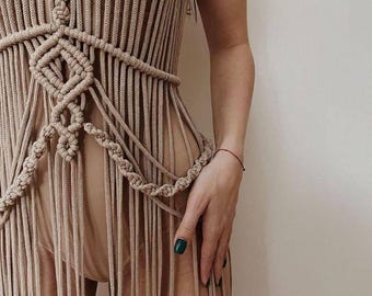 Könnte beinhalten: Beiges Makramee-Kleid mit Neckholder-Ausschnitt und Fransen. Das Kleid hat aufwendige Knoten und ein geometrisches Design um die Taille. Das Kleid ist aus einem weichen, natürlich aussehenden Material gefertigt.