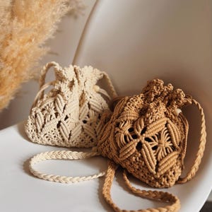Op de afbeelding: Twee macramé tassen met trekkoord in crème- en bruintinten liggen op een witte stoel. De tassen hebben ingewikkelde geweven patronen en gevlochten bandjes. De crèmekleurige tas is links, de bruine rechts. Op de achtergrond gedroogd siergras.