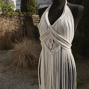 May include: A white macrame halter top with a geometric pattern and fringe. The top is displayed on a mannequin against a backdrop of greenery.