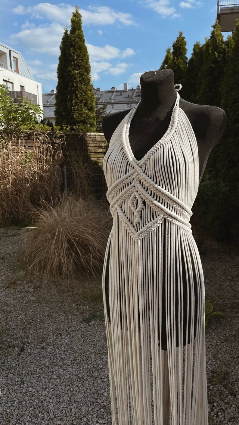 May include: A white macrame halter top with a geometric pattern and fringe. The top is displayed on a mannequin against a backdrop of greenery.