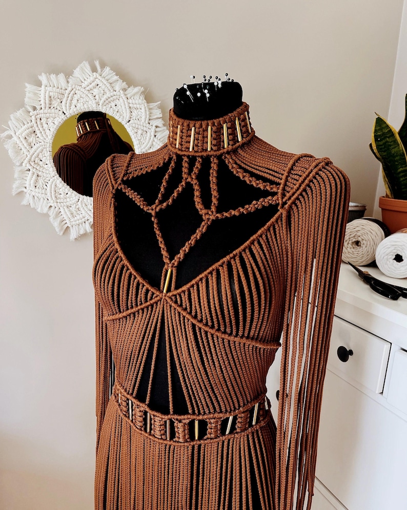 Op de afbeelding: Een bruin macram&eacute; kledingstuk met een hoge hals voorzien van goudkleurige cilindrische accenten, ingewikkelde knooppatronen op de borst, lange mouwen met franjes en een bijpassende riem met goudkleurige details, getoond op een zwarte paspop.