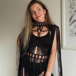 Op de afbeelding: Een zwart macram&eacute; topje met een hoge chokerhalslijn met goudkleurige accenten, een gestructureerd lijfje met verticale koorddetails en lange franjes vanaf de schouders en taille, gecombineerd met een bijpassend zwart basiskledingstuk.