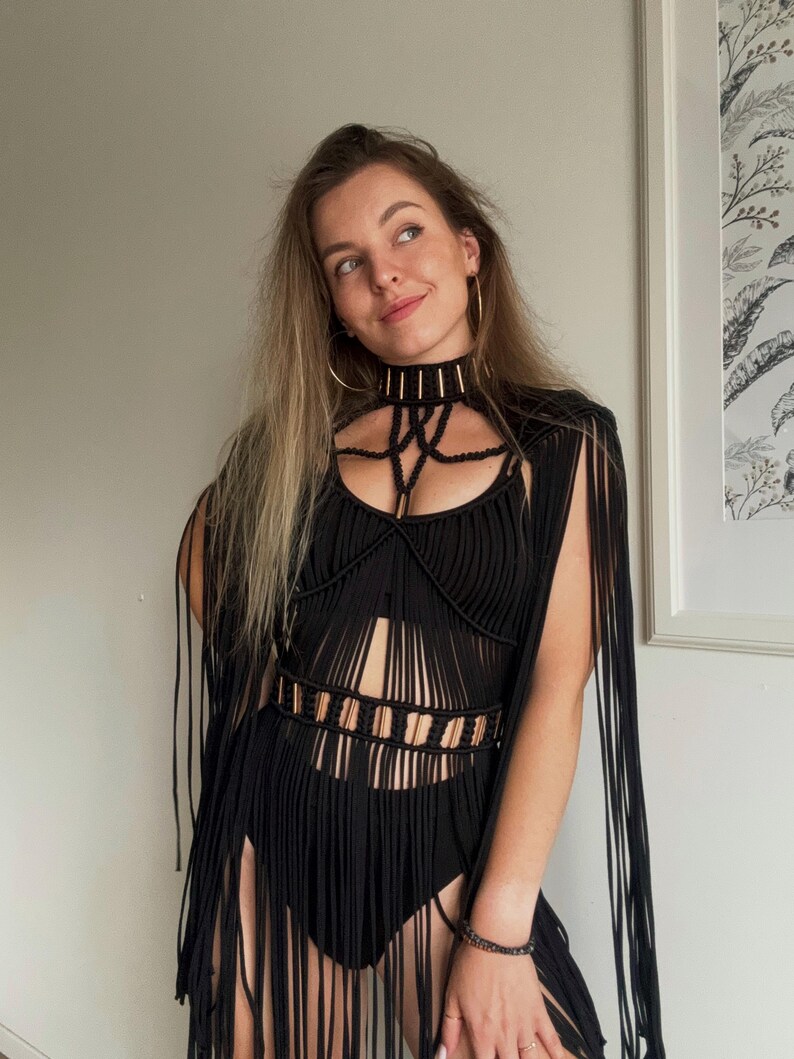 Op de afbeelding: Een zwart macram&eacute; topje met een hoge chokerhalslijn met goudkleurige accenten, een gestructureerd lijfje met verticale koorddetails en lange franjes vanaf de schouders en taille, gecombineerd met een bijpassend zwart basiskledingstuk.