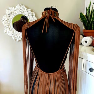 Op de afbeelding: Een bruin macram&eacute; kledingstuk met een hoge kraag met goudkleurige cilindrische accenten, een open rugontwerp en lange, vloeiende franjes die zich uitstrekken van de schouders tot aan het onderste deel van het kledingstuk.