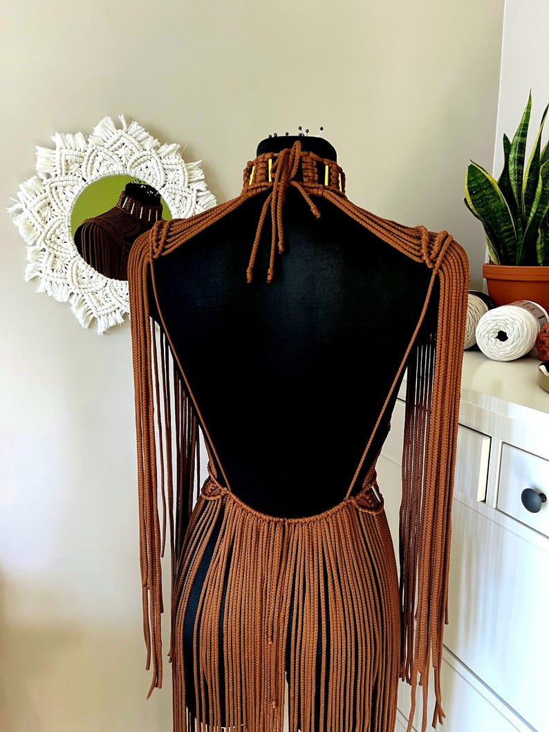 Op de afbeelding: Een bruin macram&eacute; kledingstuk met een hoge kraag met goudkleurige cilindrische accenten, een open rugontwerp en lange, vloeiende franjes die zich uitstrekken van de schouders tot aan het onderste deel van het kledingstuk.