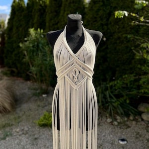 May include: A white crocheted halter top with a geometric pattern and long fringe. The top is displayed on a mannequin against a backdrop of green foliage.
