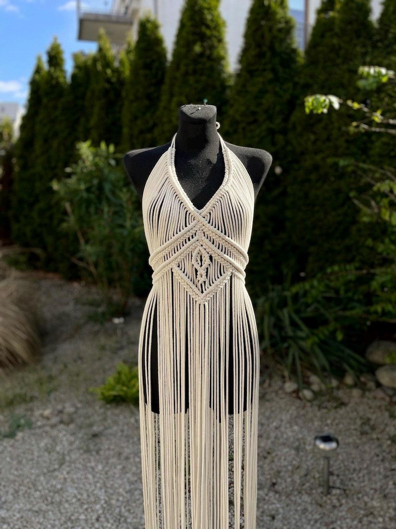 May include: A white crocheted halter top with a geometric pattern and long fringe. The top is displayed on a mannequin against a backdrop of green foliage.