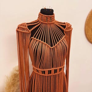 Op de afbeelding: Een terracotta-kleurig macramé kledingstuk met een hoge hals, lange mouwen en een aansluitend lijfje. Het stuk is gemaakt van ingewikkelde geknoopte koordpatronen, wat zorgt voor een gestructureerd kooi-achtig ontwerp met lange, vloeiende franjes.
