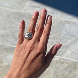Puede incluir: Un anillo de plata con una forma orgánica texturizada. El anillo se lleva en el dedo medio de una mano.