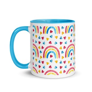 May include: White ceramic mug with a blue handle and interior. The mug features a repeating pattern of watercolor rainbows in red, yellow, and blue, interspersed with red, yellow, and blue hearts.