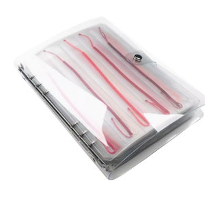 May include: A clear plastic organizer containing several red and pink knitting needles. The needles are of varying lengths and are secured within individual clear sleeves. The organizer has a silver metal binding and a snap closure.